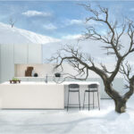 A modern white kitchen island and stools stand in a snow-covered landscape, beside a leafless tree under a clear blue sky, blending indoor elements with an outdoor winter setting.
