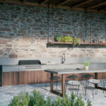 A modern outdoor kitchen features a wooden table with stools, a built-in grill, and green plants, set against a rustic stone wall under a wooden beam ceiling.