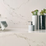 Bathroom countertop with a white marble finish; holds a light grey soap dispenser and two cylindrical vases containing succulents. A towel is casually draped nearby.