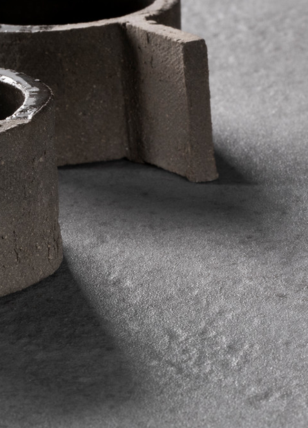 A rough, circular concrete object with protruding segments rests on a textured gray surface, casting soft shadows in a minimalist setting.
